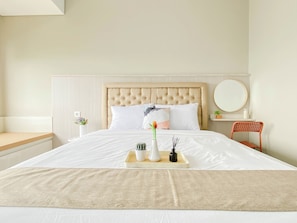 A neatly made bed with a tufted headboard, a bedside table with a vase and flowers, and a chair with a round mirror. - Bright And Cozy Studio At Royal Sentul Park Apartment (Bogor)