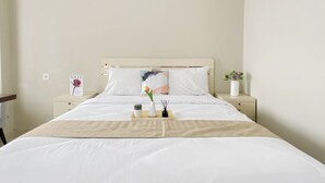 A neatly made bed with white bedding, a beige throw, and a nightstand with a vase and a picture frame. - Tranquil Studio For 2 Pax At Royal Sentul Park Apartment (Bogor)