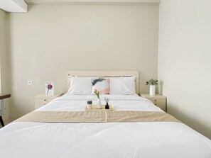 Una cama bien tendida con ropa blanca, un plaid color beige y una mesita de noche con un florero y un marco de fotos.