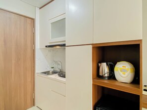 Una cocina compacta con estufa integrada, un lavamanos y una pequeña estantería con hervidor y cocedor de arroz.