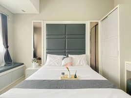 A neatly made bed with a grey headboard, a bedside table with a vase and a candle, and a window with curtains.