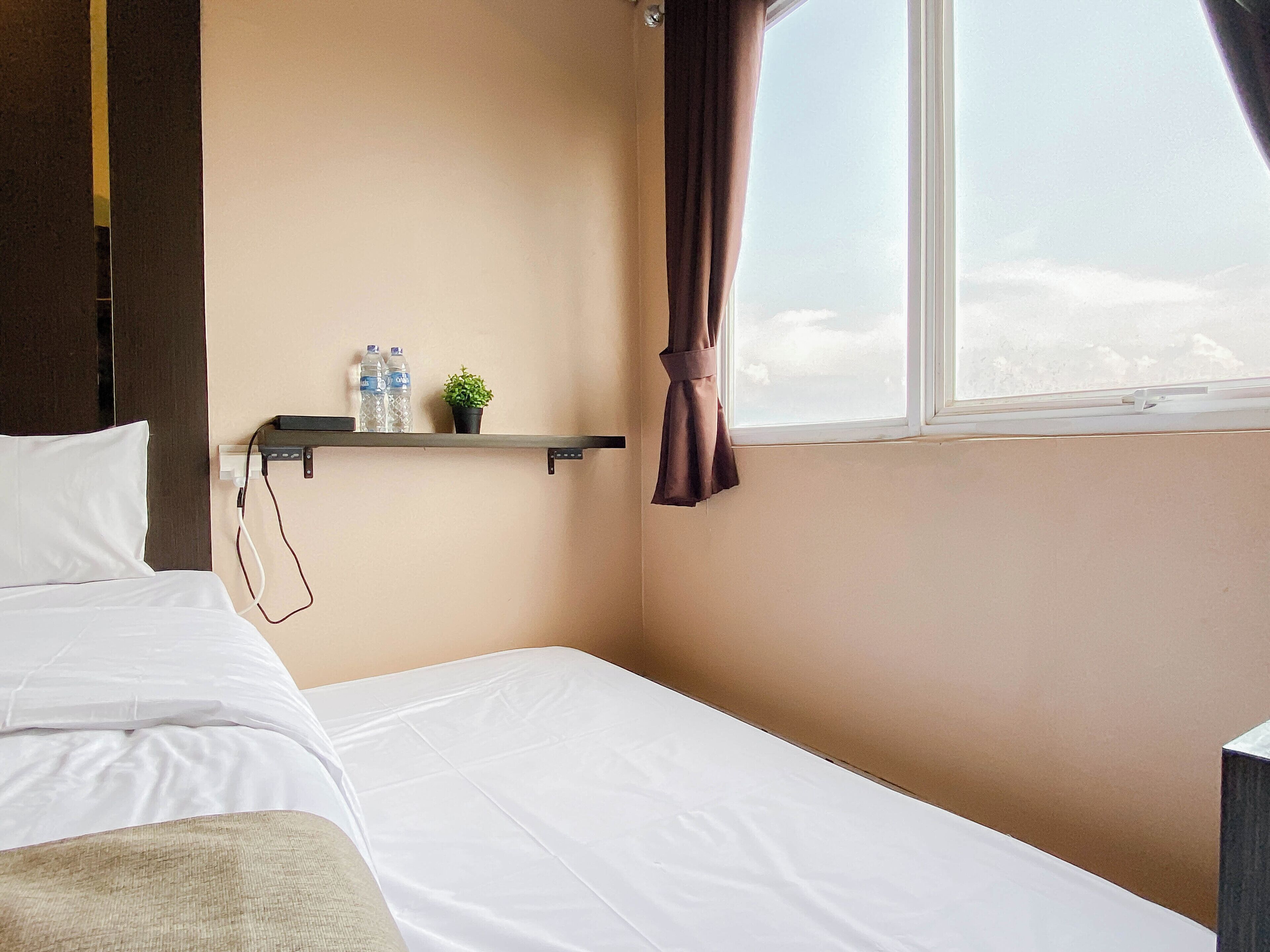 A hotel room with a bed, a shelf with a plant and water bottles, a window with curtains, and a view of the sky.