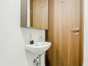 A bathroom with a white sink, a wooden door, and a mirror above the sink. - Wooden Touch Studio Room At Apartment Lrt City Sentul - Royal Sentul Park (Bogor)