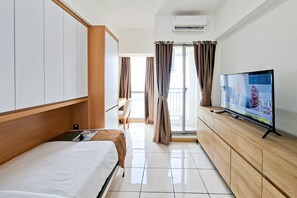 A hotel room with a bed, a TV, a wooden cabinet, and a view through a glass door. - Cozy And Decent Studio Room At Apartment M-Town Residence (Gading Serpong)