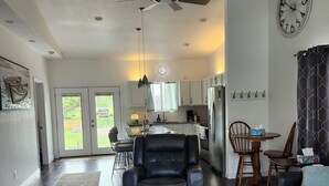 A spacious living area with a ceiling fan, a dining table with chairs, a kitchen area, and a large clock on the wall.