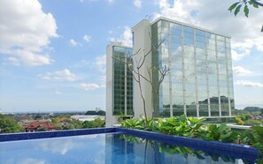 A modern building with a glass facade, a swimming pool with blue tiles, and surrounding greenery. - Best Deal Studio At Mataram City Apartment (Ngaglik)