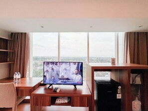 A hotel room with a large TV on a wooden stand, a small table with a remote, a bottle of water, and a bookshelf. - Best Deal Studio At Mataram City Apartment (Ngaglik)