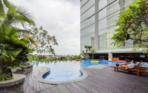 A swimming pool with a wooden deck, surrounded by greenery and a modern building. - Good Deal Studio Apartment At Mataram City (Yogyakarta)