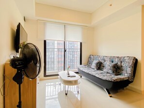 A modern living room with a sofa, a small table, a standing fan, and a TV mounted on the wall.