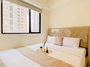 A hotel room with a bed, a window with blinds, a bedside table with a tray, and a view of buildings outside. - Cozy 3Br Apartment With Minimalist Style At Meikarta (Cikarang)