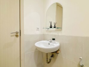 Un bagno con un lavandino bianco, uno specchio, uno scaffale in vetro con articoli da toilette e una porta.