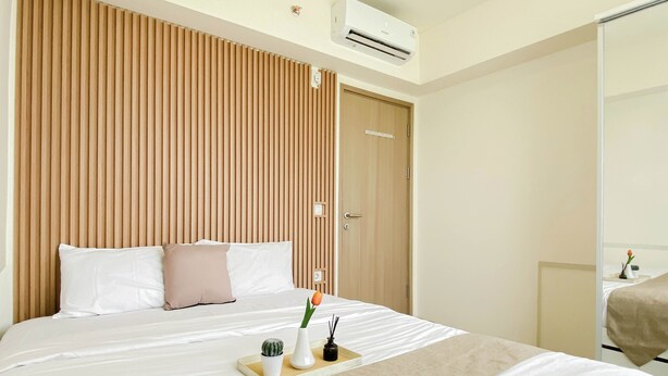 A hotel room with a bed, a wooden headboard, an air conditioning unit, a door, and a small table with a plant and a tray. - Restful And Spacious 3Br At Apartment Meikarta District 1 (Cikarang)