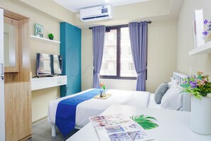 Ein Hotelzimmer mit Bett, Schreibtisch mit Computer, Fenster mit Vorhängen, Regal mit Büchern und Pflanze sowie eine Wand-Klimaanlage.