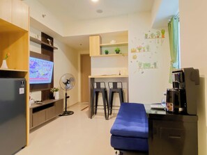 A compact living space with a kitchenette, a TV, a fan, a sofa, and a coffee machine. - Wooden Accents 2Br For 3 Pax At Meikarta District 2 Apartment (Cikarang)
