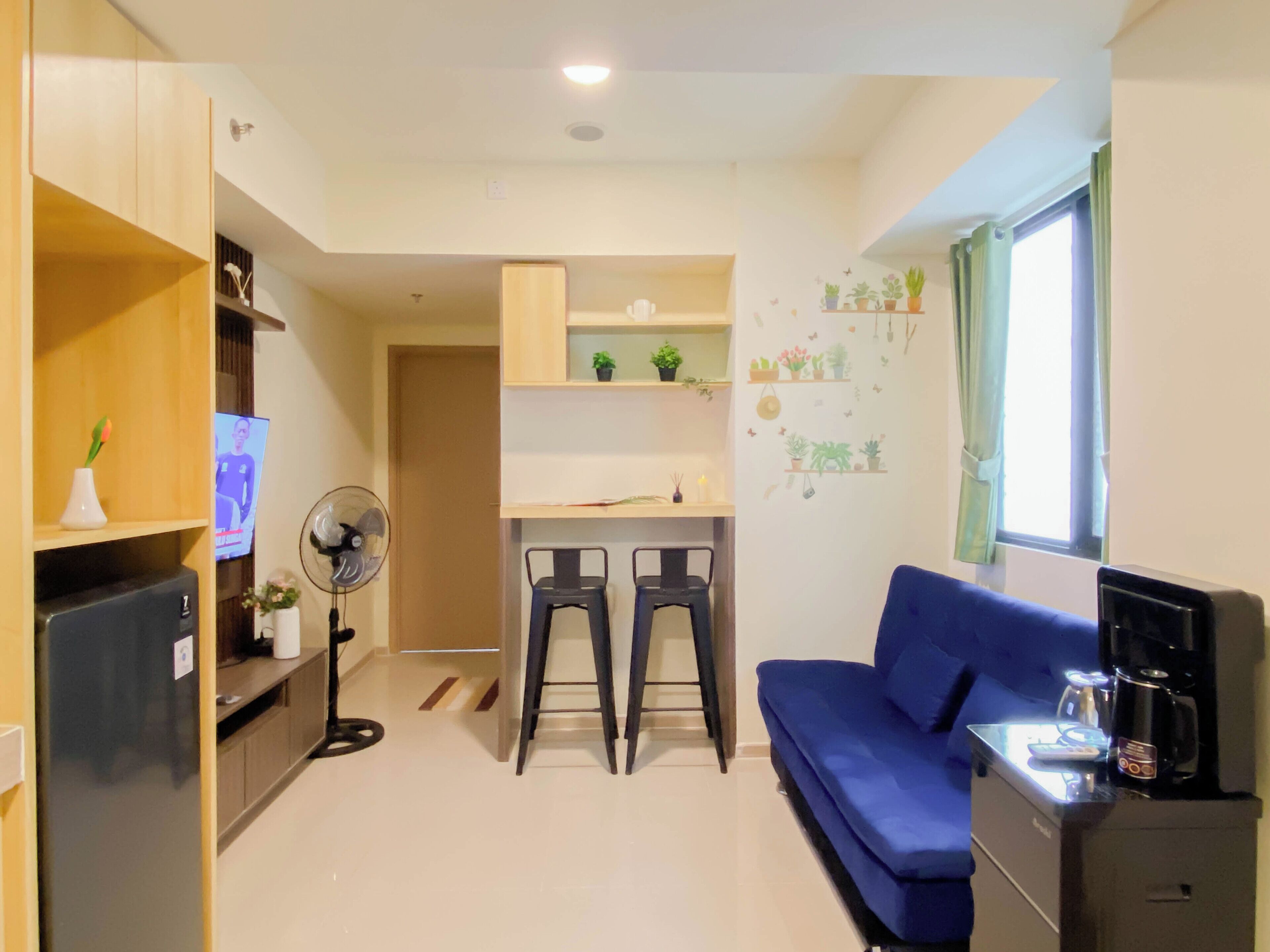 A compact living space with a kitchenette, a sofa, two chairs, a TV, a fan, and a coffee machine.