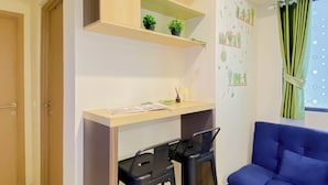 A room with a blue sofa, a wooden desk with a chair, a small shelf with plants, and a wall with potted plant decorations. - Wooden Accents 2Br For 3 Pax At Meikarta District 2 Apartment (Cikarang)