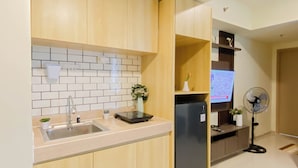 A modern kitchen with wooden cabinets, a tiled backsplash, a stainless steel sink, a black refrigerator, and a small fan. - Wooden Accents 2Br For 3 Pax At Meikarta District 2 Apartment (Cikarang)