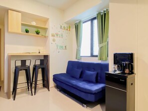 Una habitación con un sofá azul, dos sillas negras, una mesita, una cafetera y una ventana con cortinas.