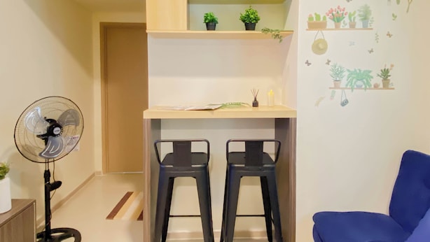 A compact living space with a kitchenette, two bar stools, a standing fan, and a blue sofa. - Wooden Accents 2Br For 3 Pax At Meikarta District 2 Apartment (Cikarang)