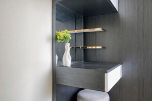 A modern interior with a dark wood panel wall, a white vase with green foliage, and a minimalist shelf unit. - Serene Look 2Br Apartment Meikarta District 2 (Cikarang)