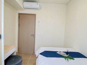 A small, clean room with a bed, a toilet, a wooden door, and a wall-mounted air conditioner. - Modern Versatile 3Br At Meikarta District 2 Apartment (Cikarang)