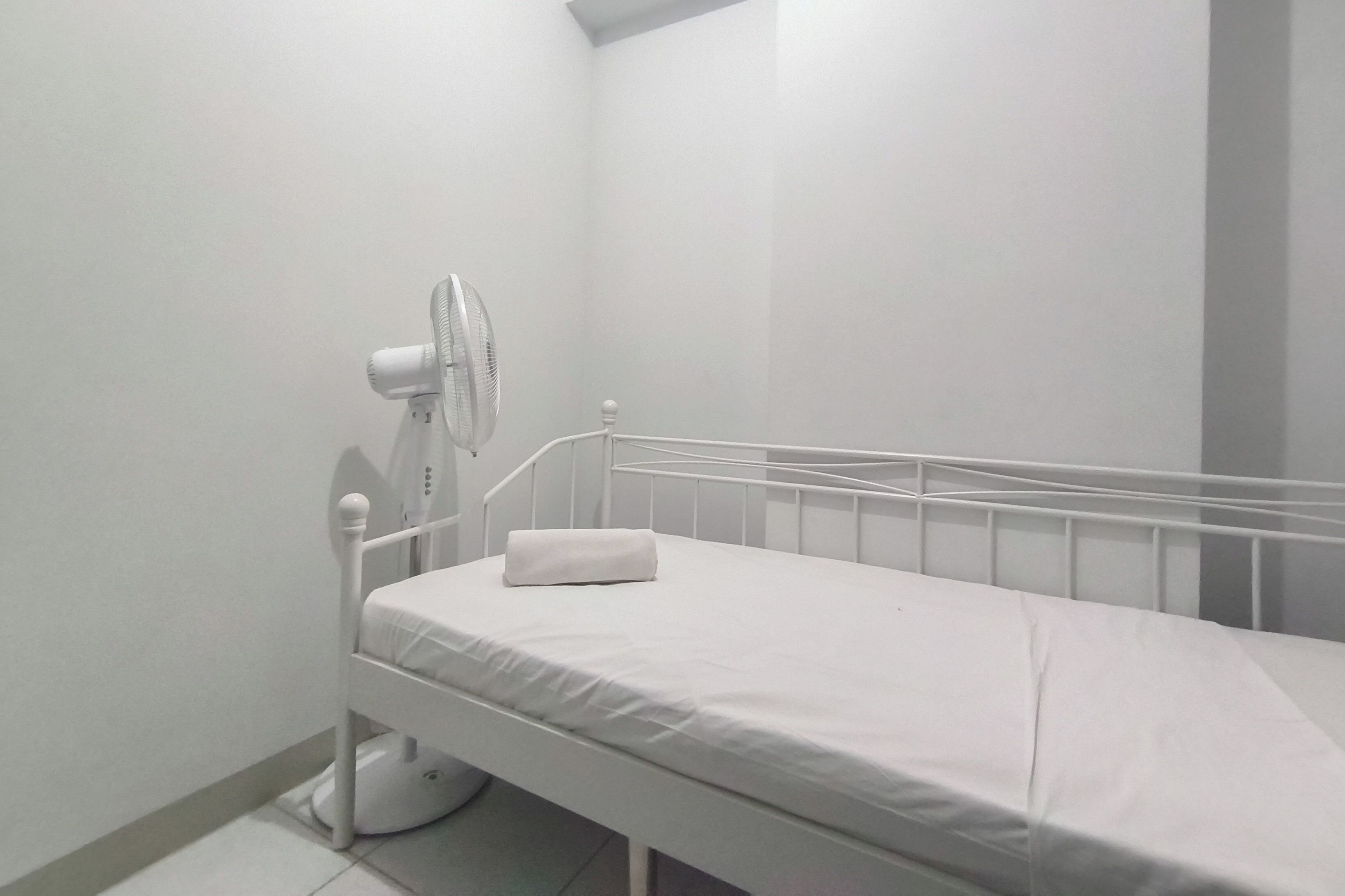 A small, white, single-bed room with a white fan and a white pillow on the bed.