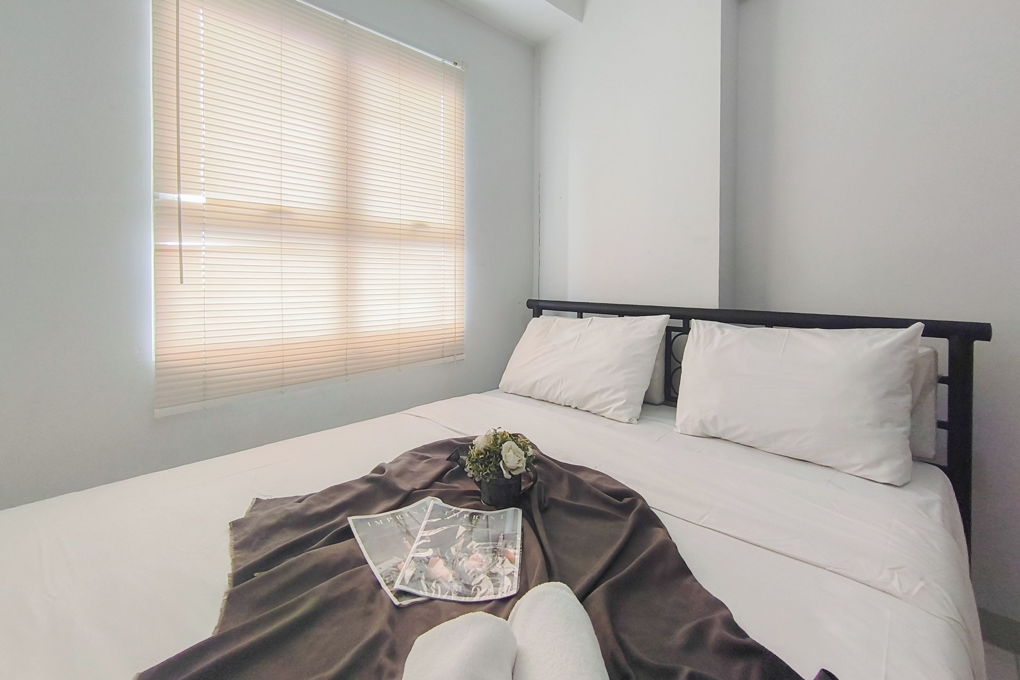 Een slaapkamer met een bed, witte kussens, een bruine deken, een tijdschrift en een kleine plant.