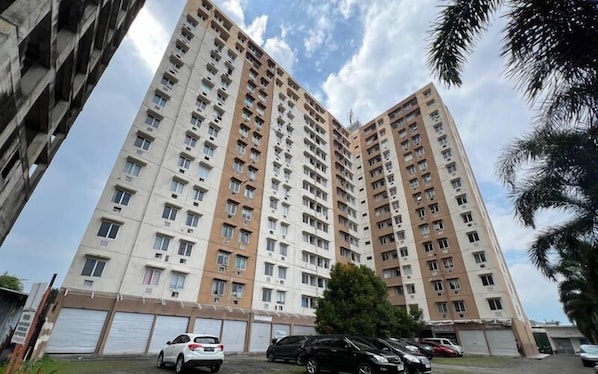 A high-rise residential building with multiple floors, large windows, and a parking area in front.