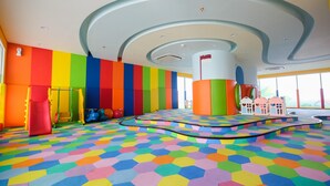 Un área de juegos colorida con piso de patrón hexagonal, un pilar central grande y vistas al exterior a través de ventanas amplias.
