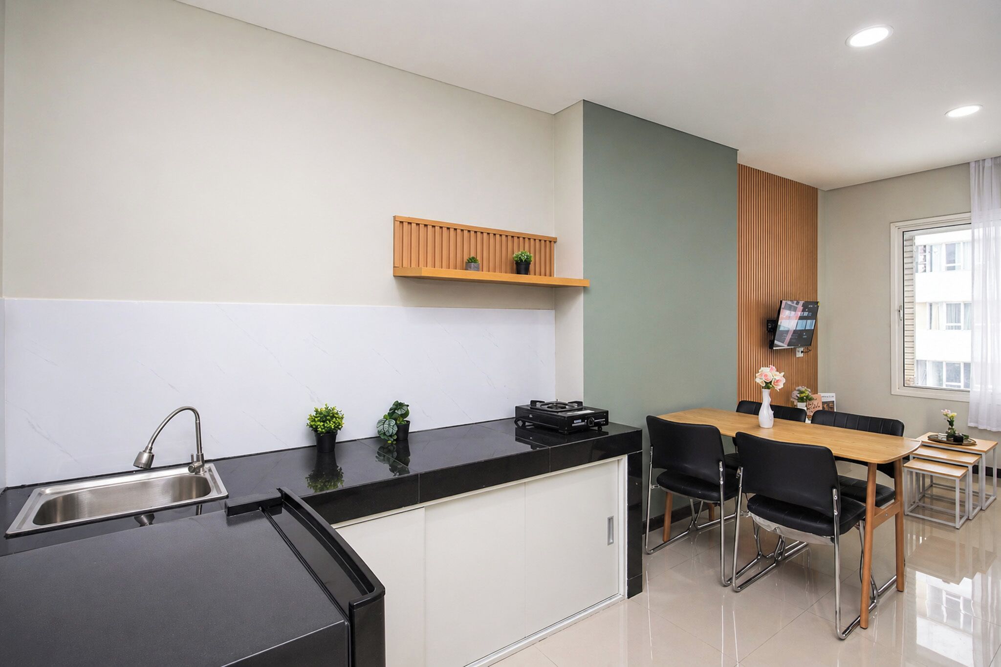 Una cocina moderna con un lavamanos empotrado, una estantería de madera con plantas, una zona de comedor con mesa y sillas, y un televisor montado en la pared.