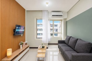 Ein modernes Wohnzimmer mit Flachbildfernseher, einem grauen Sofa, einer Wandpaneele aus Holz, einem Fenster mit Blick auf Gebäude und einer Klimaanlage.