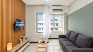 A modern living room with a flat-screen TV, a grey sofa, a wooden panel wall, a window with a view of buildings, and an air conditioning unit. - Functional Designed 2Br Apartment At Nifarro Park (Jakarta)