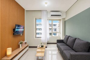 Un salon moderne doté d’un téléviseur à écran plat, d’un canapé gris, d’un mur recouvert de panneaux en bois, d’une fenêtre donnant sur des immeubles et d’un appareil de climatisation.