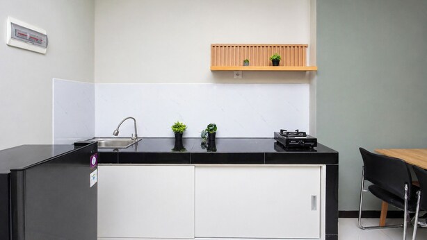 A modern kitchen with a black countertop, white cabinets, a wooden shelf with plants, and a small sink. - Functional Designed 2Br Apartment At Nifarro Park (Jakarta)