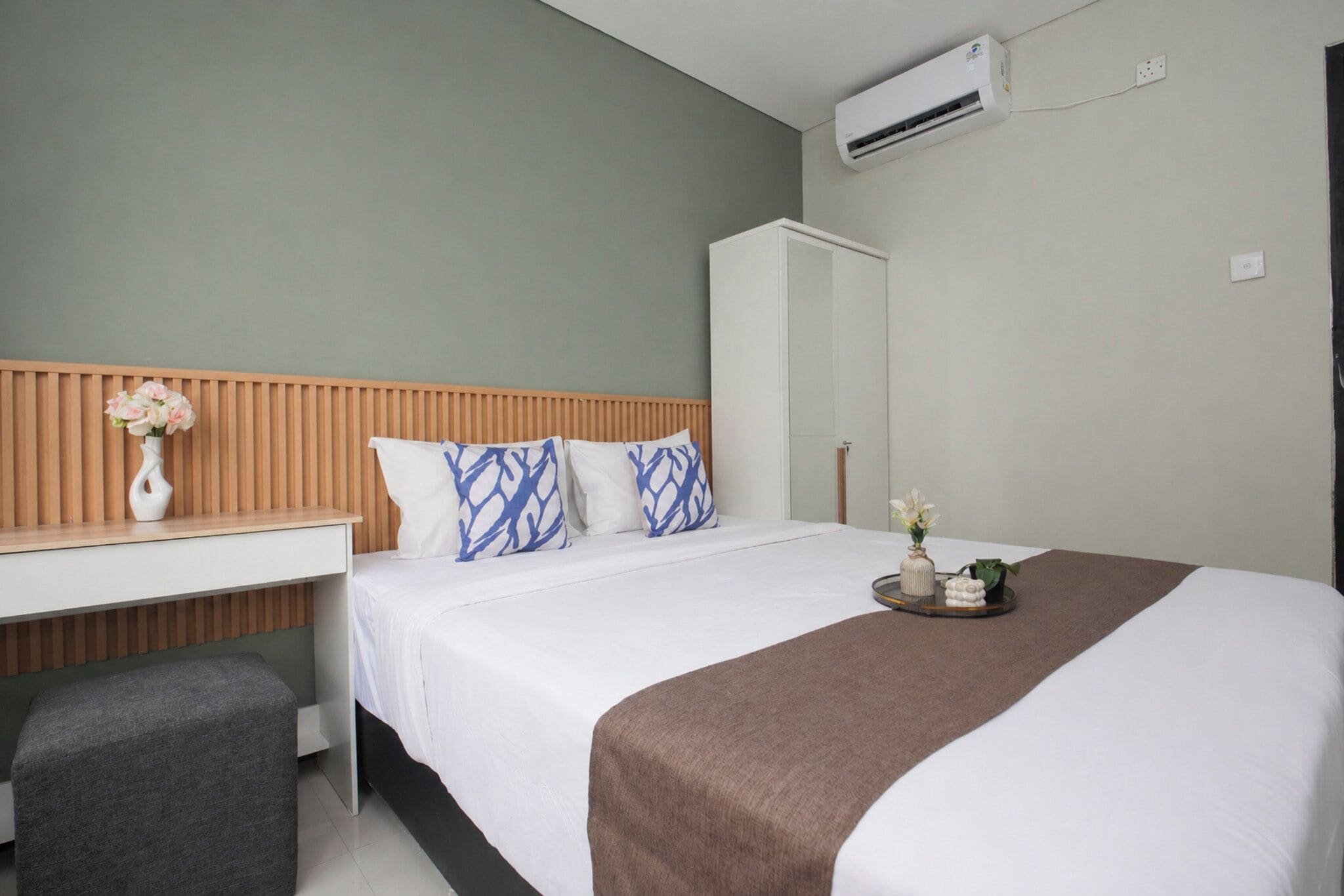 A hotel room with a bed, a wooden headboard, a white bedside table with a vase, a grey bench, a brown throw blanket, and a tray with flowers.