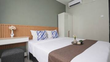 A hotel room with a bed, a wooden headboard, a white bedside table with a vase, a grey bench, a brown throw blanket, and a tray with flowers.