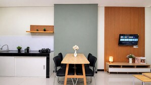 A modern kitchen with a wooden table, black chairs, a TV mounted on a wooden panel, and a small shelf with plants. - Functional Designed 2Br Apartment At Nifarro Park (Jakarta)