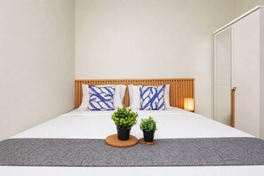 A neatly made bed with a wooden headboard, two pillows with a blue pattern, and two potted plants on a grey rug. - Cozy Living 2Br Nifarro Park Apartment (Jakarta)