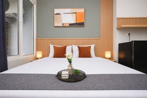 A hotel room with a bed, a wall-mounted abstract painting, a wooden headboard, a side table with a lamp, and a black refrigerator. - Serene Sage Touch Studio Room At Apartment Nifarro Park (Jakarta)