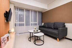A modern room with a grey sofa, two small round tables, a TV mounted on a wooden panel, and a large window with sheer curtains. - Roomy Urban Living In 3Br At Apartment Nifarro Park (Jakarta)