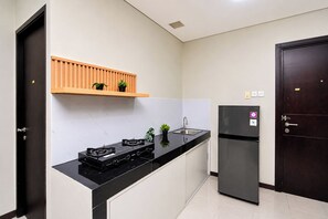 A compact kitchen with a black countertop, a built-in stove, a refrigerator, and a wooden shelf above the sink. - Roomy Urban Living In 3Br At Apartment Nifarro Park (Jakarta)