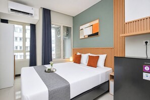 Ett hotellrum med en säng, ett skrivbord, en luftkonditionering, ett fönster med gardiner och ett konstverk som hänger på väggen.