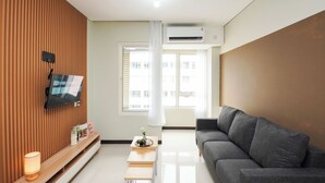 Een moderne woonkamer met een flatscreen televisie aan een houten wand, een grijze bank, een klein houten tafeltje met een lamp en een raam met witte gordijnen.