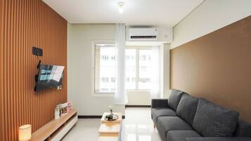 A modern living room with a flat-screen TV mounted on a wooden wall, a grey sofa, a small wooden table with a lamp, and a window with white curtains.