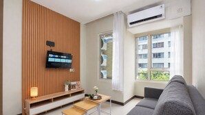 Un salón moderno con un televisor de pantalla plana montado en una pared de paneles de madera, un sofá gris, una mesita de madera y una ventana con vistas a un edificio.