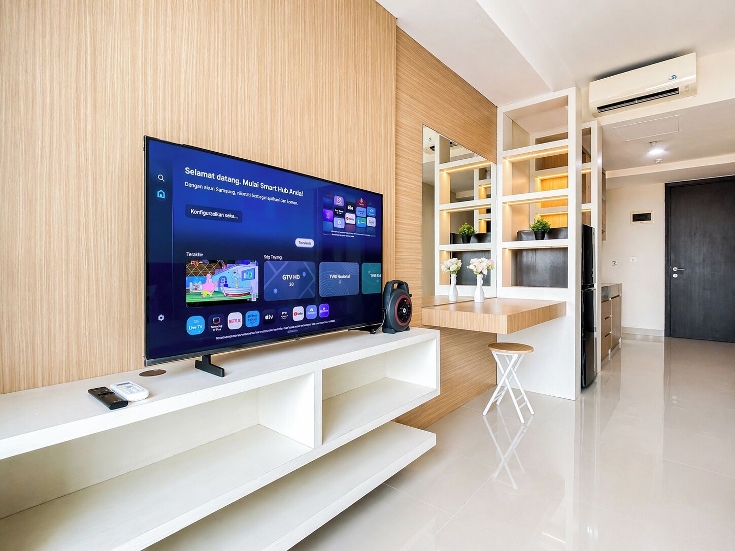 Una sala de estar moderna con un televisor de pantalla plana grande montado en una pared de madera, un mueble de entretenimiento blanco debajo y un taburete blanco pequeño.