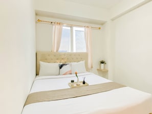 Ein ordentlich bezogenes Bett mit Kopfteil, ein Nachttisch mit Vase und Pflänzchen, und ein Fenster mit Vorhängen.
