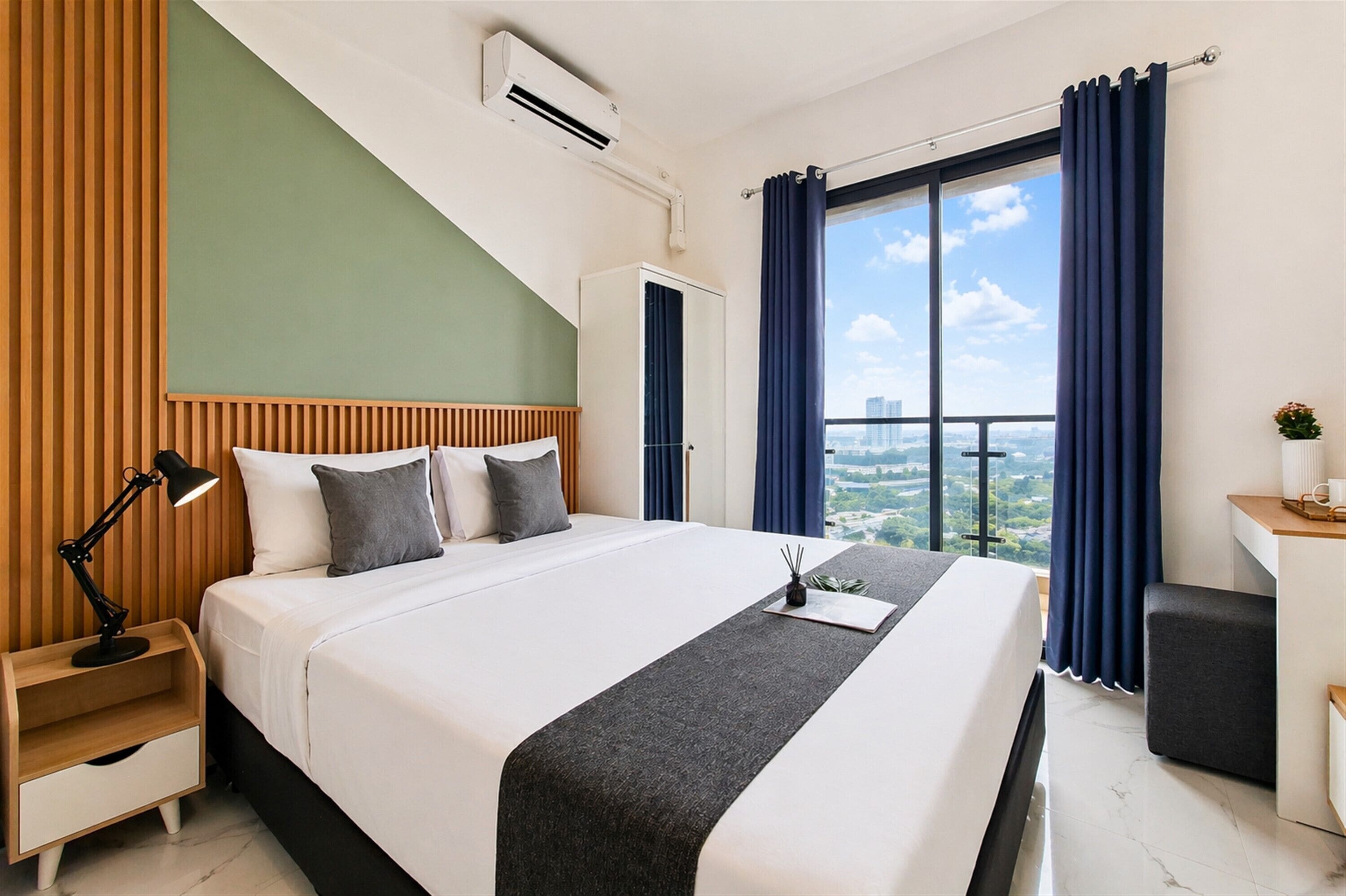 Ein Hotelzimmer mit Bett, Nachttischlampe, Klimaanlage und Blick auf eine Stadtlandschaft durch große Fenster.