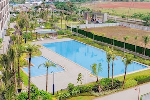 Ein großer Swimmingpool, umgeben von Palmen und viel Grün, mit einem Sportplatz im Hintergrund.