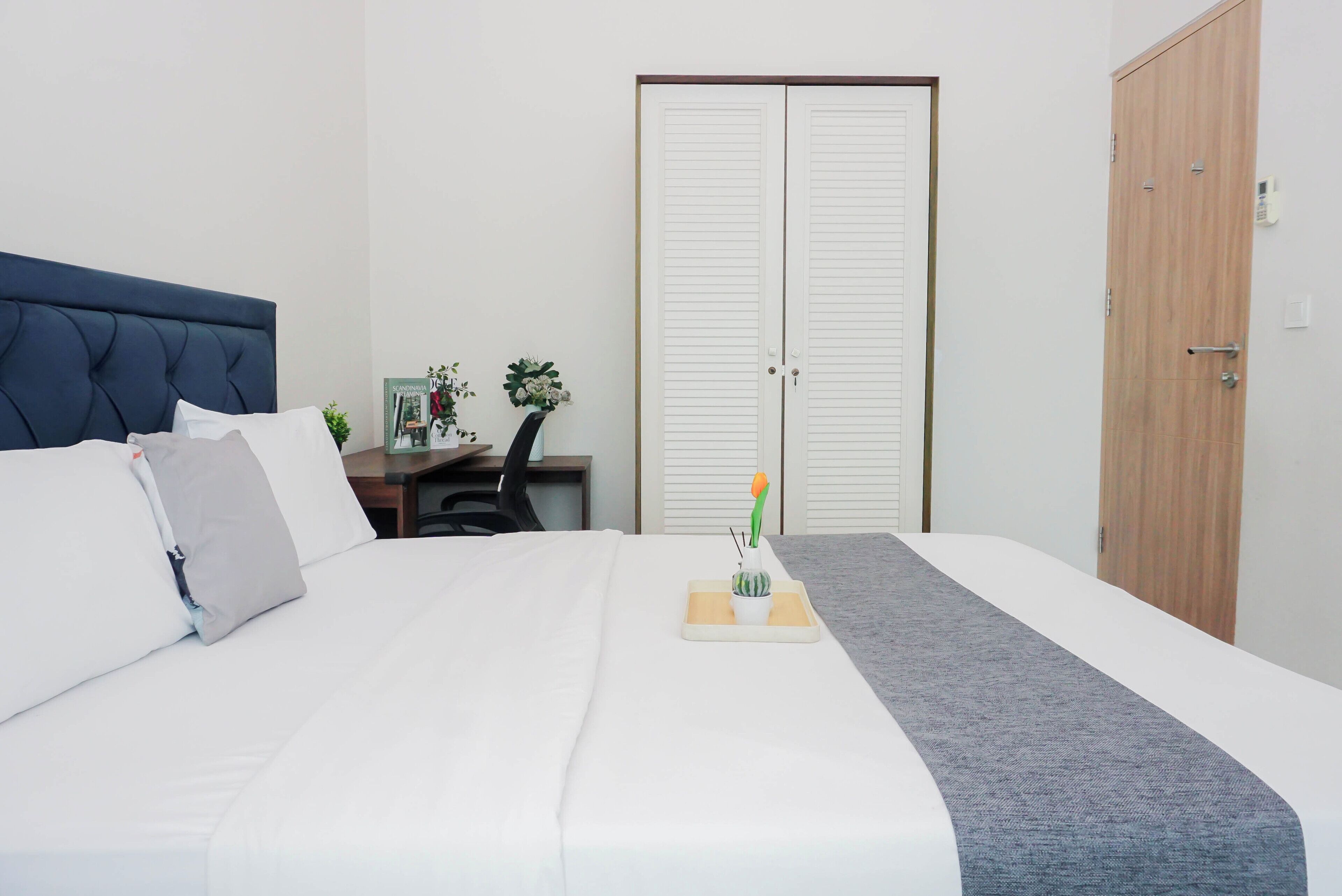 Una cama bien tendida con ropa blanca, un plaid gris y un pequeño cactus en una bandeja. Se observa una puerta de madera y un armario blanco al fondo.